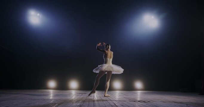 Back view on professional female diligent ballet dancer practicing dance in white dress in the studio in blue spotlight. Rear of young graceful hard-working ballerina dancing elements of ballet.