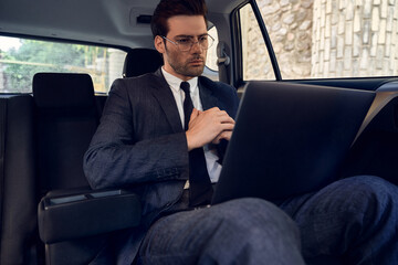 Confident young businessman seriously and thoughtfully looks at the laptop while sitting in the car.