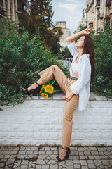 Young happy beautiful girl is walking in a shirt and pants with a bouquet of flowers of sunflowers on a city street