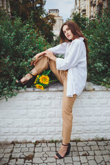 Young happy beautiful girl is walking in a shirt and pants with a bouquet of flowers of sunflowers on a city street