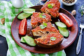 Eggplant stuffed with minced chicken, cheese and raisins in tomato sauce on a brown clay plate on a black concrete background.
