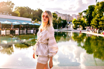 Sunny summer image of pretty stylish elegant blonde woman posing at city park in Europe, positive mood, walking alone.