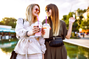 Two happy pretty best friends girls spending time at city park, cheers tasty milkshakes, focus on hands and sweet drinks.