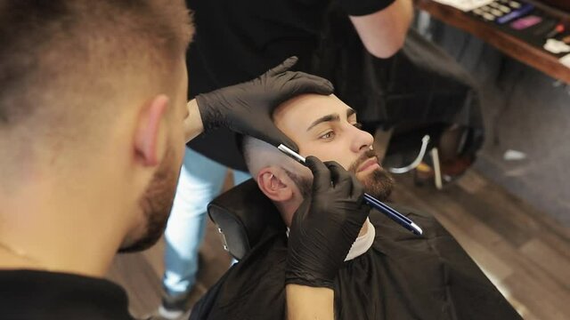 Hands in black gloves shaving the face of a man. Professional barber shaves customer beard with straight razor. Beard cut with old-fashioned blade at barbershop.