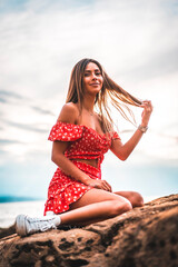 A young brunette Caucasian woman in a red dress on the beach of Itzurrun in the town of Zumaia, Gipuzkoa. Basque Country. Lifestyle session, on a rock smiling sitting with the sun behind