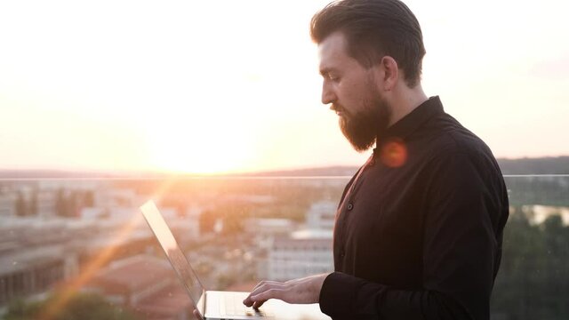 Attractive Stylish Businessman Dressend In Black Shirt Typing On Keyboard Writing A Message On His Computer Laptop Staying In Beautiful Terrace On Roof Of A Building. Beauty Cityscape On Background.