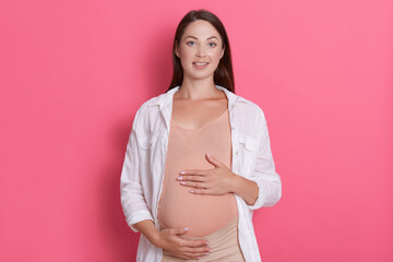 Adorable happy young pregnant woman touching belly and looking directly at camera while posing isolated over pink background, expectant mother wearing white shirt and bodysuit.