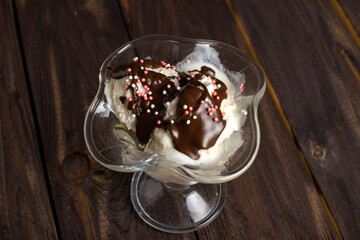 White balls of vanilla ice cream poured with chocolate topping in a dessert glass. Decorated ice cream on an old table made of rough wood. Copyspace.