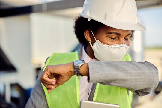 Black civil engineer with face mask sneezing into elbow outdoors.