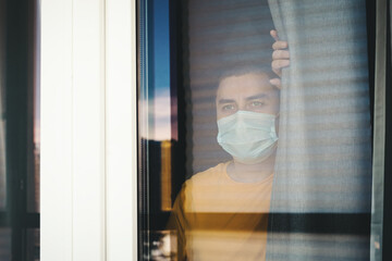  Quarantine man in medical mask on face  looking through the window.