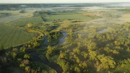 Tuinposter Aerial view of rural landscape with lush greenery, trees, river in the foreground and farmhouse, farmland in the background. American midwestern countryside scenery in the early sunny morning, summer © alenamozhjer