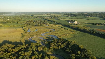 Aerial view of rural landscape with lush greenery, trees, river in the foreground and farmhouse, farmland in the background. American midwestern countryside scenery in the early sunny morning, summer © alenamozhjer