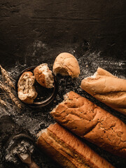 Broken bread baguettes on black slate background with wheat spikelets, sackcloth, flour. Pastries and bread in a bakery, Food concept, top view