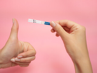 Female hand holding positive pregnancy test and Showing thumb up gesture isolated on pink background. The abbreviation HCG on the blue bar means Human chorionic gonadotropin is a hormone produced by