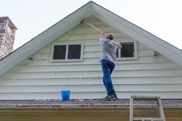 Man stands on porch roof to wash house with brush on extension
