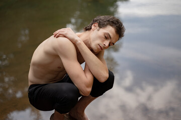 Shirtless man with eyes closed crouching on rock against lake in forest