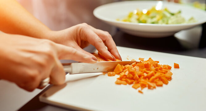 Woman cuts into small cubes on a plank carrots for salad, which stands against the background in a plate - Powered by Adobe