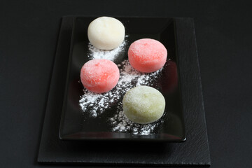 Four types of Japanese dessert mochi - pomegranate with honey, green matcha tea, strawberry, coconut on a black plate on a black table.
