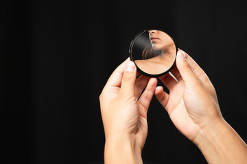 Hispanic woman looking at herself in a mirror with black background - close up hands holding a small mirror with the reflection of a face in it - reflection of her neck and face