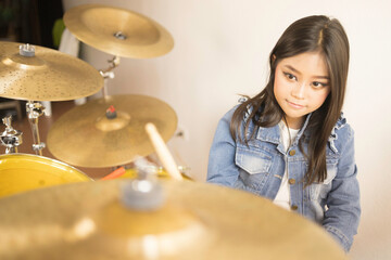 A cute Asian elementary school girl with long hair and a denim jacket happily playing drums in a music classroom.