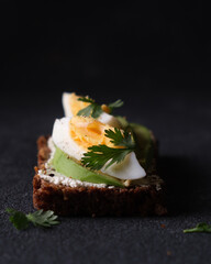 Rye bread appetizer with curd cheese, egg and avocado on black background.