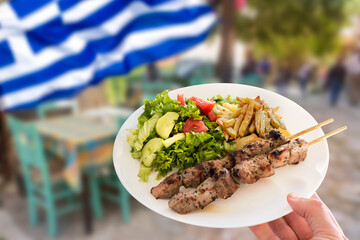 Hand holding a traditional greek pork souvlaki plate with fried potatoes and green salad. Greek tourism concept.