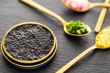 Closeup on high quality real black sturgeon caviar in a metal golden tin can aside golden spoons with pickles, red onions, egg, and herbs on a slate background. Side view.