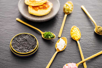 High quality real black sturgeon caviar in a metal golden tin can aside blinis in a small stone plate and golden spoons with pickles, red onions, egg, and herbs on a slate background. Side view.