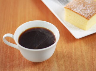 A white coffee cup with hot steam floating on the table. In the morning, there was a cake, egg, and sugar cake on the white plate for eating together.