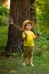 Funny kid in a hat with a cucumber and badminton on a green natural background. The concept of holidays, weekends and childhood.