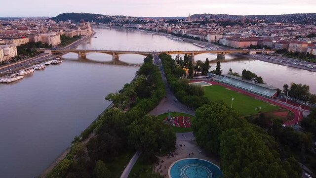 Aerial flight above margaret island with soccer field and bridge over the danube river during sunset