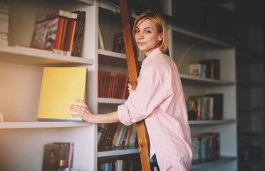 Young creative female designer picking a book to bookshelf while standing on wooden ladder at her home, blond hair woman working in apartment,girl architect holding modern book of her collection,flare