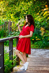 Naklejka premium a young happy pregnant woman in a red dress with flowers in her hands is resting and enjoying life in nature