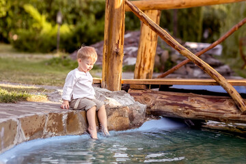A little boy is sitting on the Bank of the river. Looks at the water. Swinging his legs in the water. Enjoys a walk in nature.