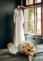 Bouquet and shoes of the bride on a table on the background of a wedding dress hanging on the window.