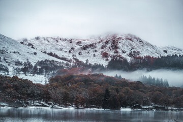 Stunning reflective, glossy lake with snow covered mountains with misty tops and autumnal forest trees in the background