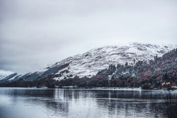 Stunning reflective, glossy lake with snow covered mountains with misty tops and autumnal forest trees in the background
