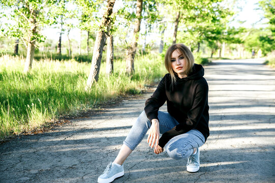 The image of a beautiful sports woman in a black hoodie squatting posing on the road.