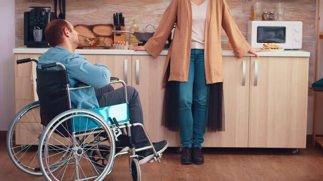 Disabled man in wheelchair having a conversation with wife in kitchen while preparing food. Guy with paralysis handicap disability handicapped difficulties getting help for mobility from love and