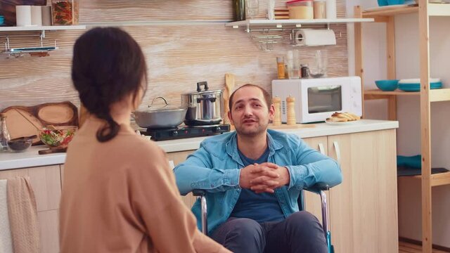 Portrait of happy disabled man in wheelchair looking at wife in kitchen. Guy with paralysis handicap disability handicapped difficulties getting help for mobility from love and relationship