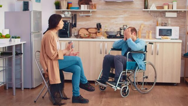 Handicapped man in wheelchair having a dispute with wife in kitchen. Guy with paralysis handicap disability handicapped difficulties getting help for mobility from love and relationship