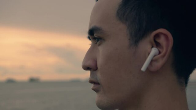 Close up of portrait handsome athlete wearing earphones listening to music while standing on the beach to looking at the ocean view beautiful sunset in summer.