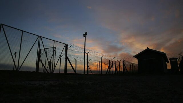 Time-lapse of a sunset  and clouds moving with prison in background silhouette with barbed wire and fence. 