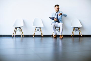 Young man sitting in the waiting room with a folder in hand and checking time before an interview