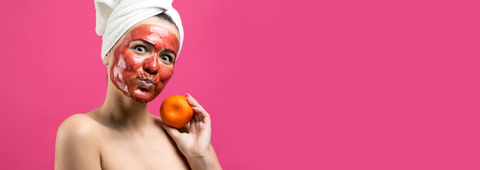 Beauty portrait of woman in white towel on head with red nourishing mask on face. Skincare cleansing eco organic cosmetic spa relax concept. A girl stands with her back holding an orange mandarin.