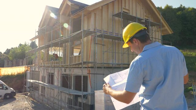 SLOW MOTION, CLOSE UP, LENS FLARE: Unrecognizable construction site manager looks at the floor plans while standing outside of a modern CLT house under construction in the countryside on a sunny day.