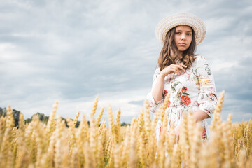 portrait of a young girl with blond long hair with a happy smile on her face, looks straight, straightens her hat, wears a light dress and a light hat, standing in a field or meadow in summer or autum