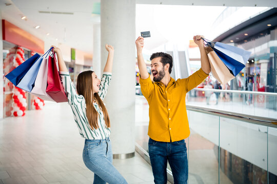 Photo of two people cheerful pretty lady handsome guy couple enjoy free time hold many bags walk shopping center raise hands use credit card discounts wear casual jeans shirt outfit indoors