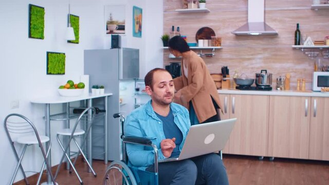 Disabled man in wheelchair using laptop in kitchen and wife is standing next to him. Corporate man with paralysis handicap disability handicapped difficulties working after accident having internet