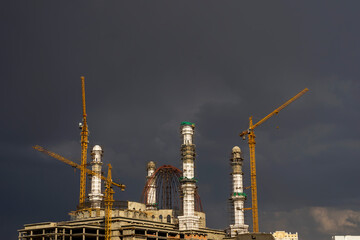 Fototapeta premium Construction of a mosque. Thunderclouds. The unfinished white mosque shines amid storm clouds. Clouds. Rainbow and Mosque. Crane. Concrete blocks and metal frame. Major construction in the city